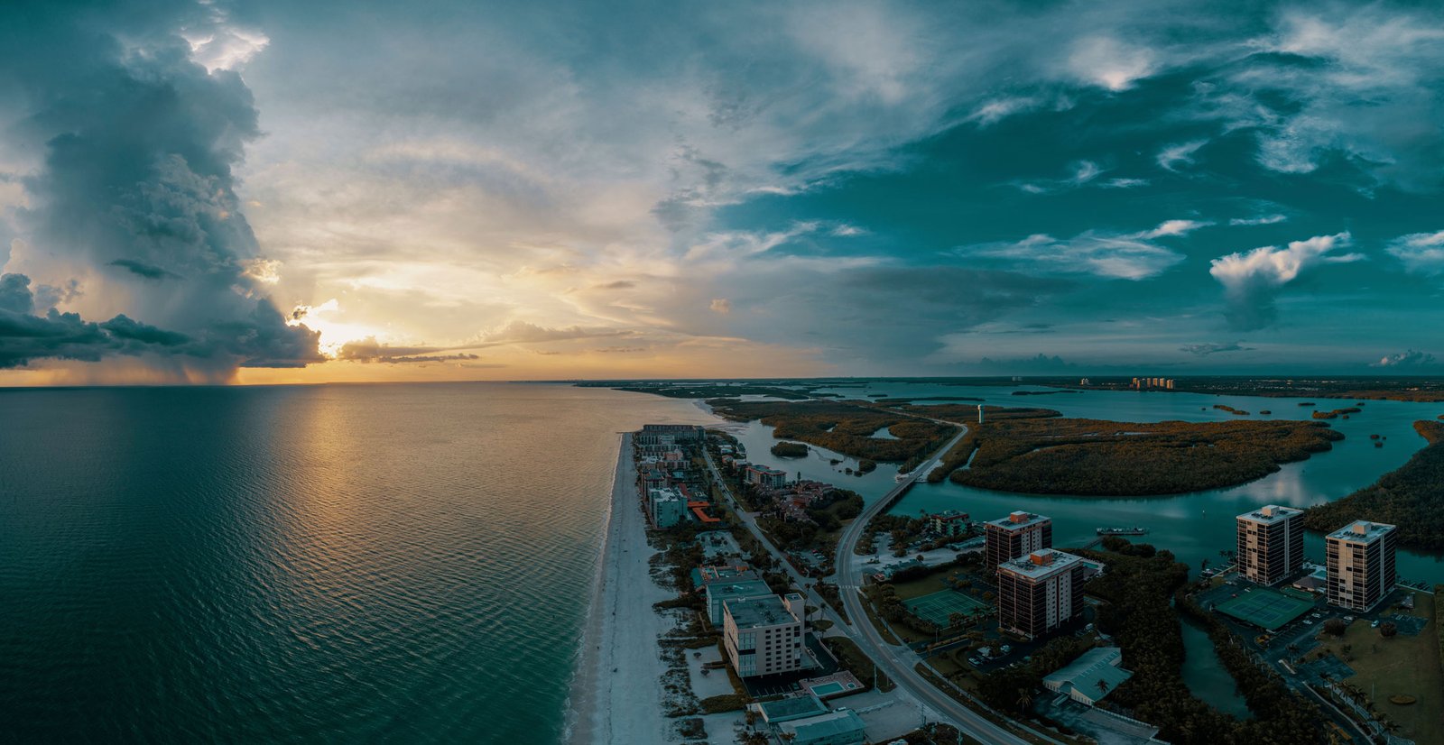 Aerial coastal cityscape at sunset in Bonita Springs, Florida, with a waterfront skyline, harbor, and dramatic clouds.